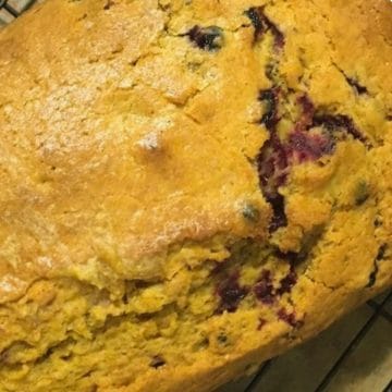 A close-up of a golden-brown loaf of bread with visible blueberries baked into it, resting on a wire cooling rack.