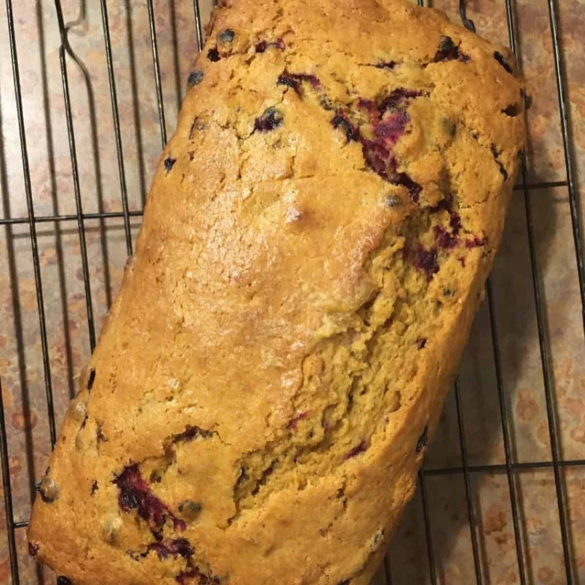 A loaf of golden-brown cranberry orange loaf quick bread with visible berry inclusions cooling on a wire rack.