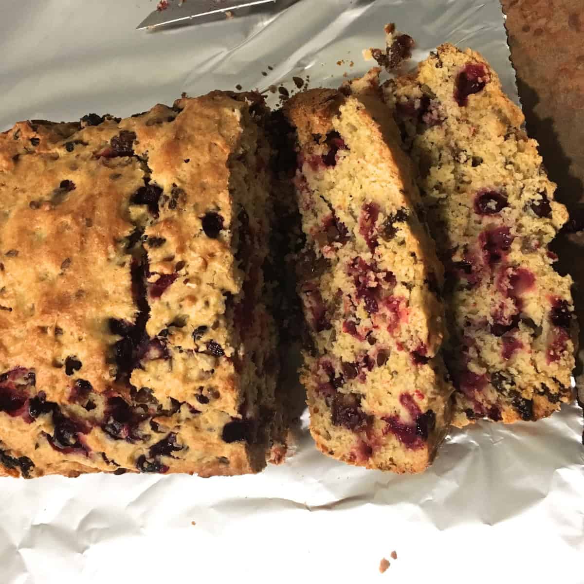 A loaf of cranberry bread partially sliced on a sheet of aluminum foil, showing a dense texture with visible cranberries throughout.