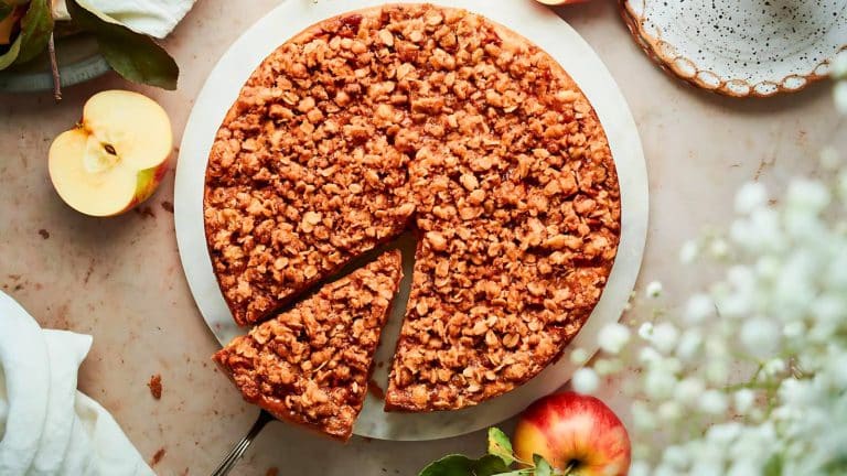 A round apple crumble cake on a white plate with a slice served. Surrounding the cake are whole and halved apples, and a couple of white flowers.