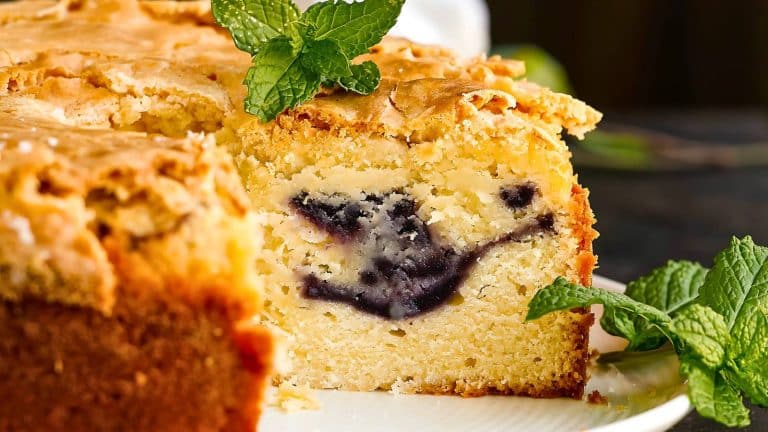 A sliced piece of berry cake with a golden crust and visible berry filling. Mint leaves are garnishing the cake, placed on a white plate.