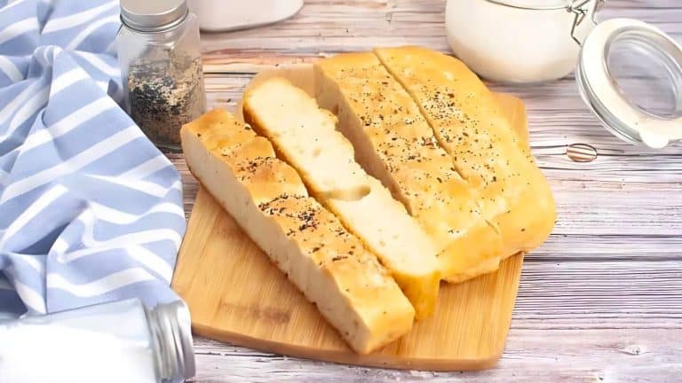 Sliced focaccia bread with herbs on a wooden cutting board, next to a striped cloth, a jar of seeds, and a canister.