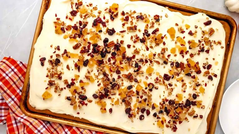 A frosted sheet cake topped with chopped nuts and dried fruit on a baking tray, next to a red checkered cloth.