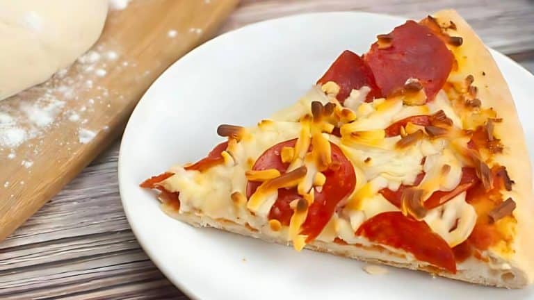 A slice of pepperoni pizza with melted cheese on a white plate. Dough and flour are visible in the background.