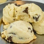 Four chocolate chip cookies are stacked on a silver plate, with one cookie showing a bite taken out.