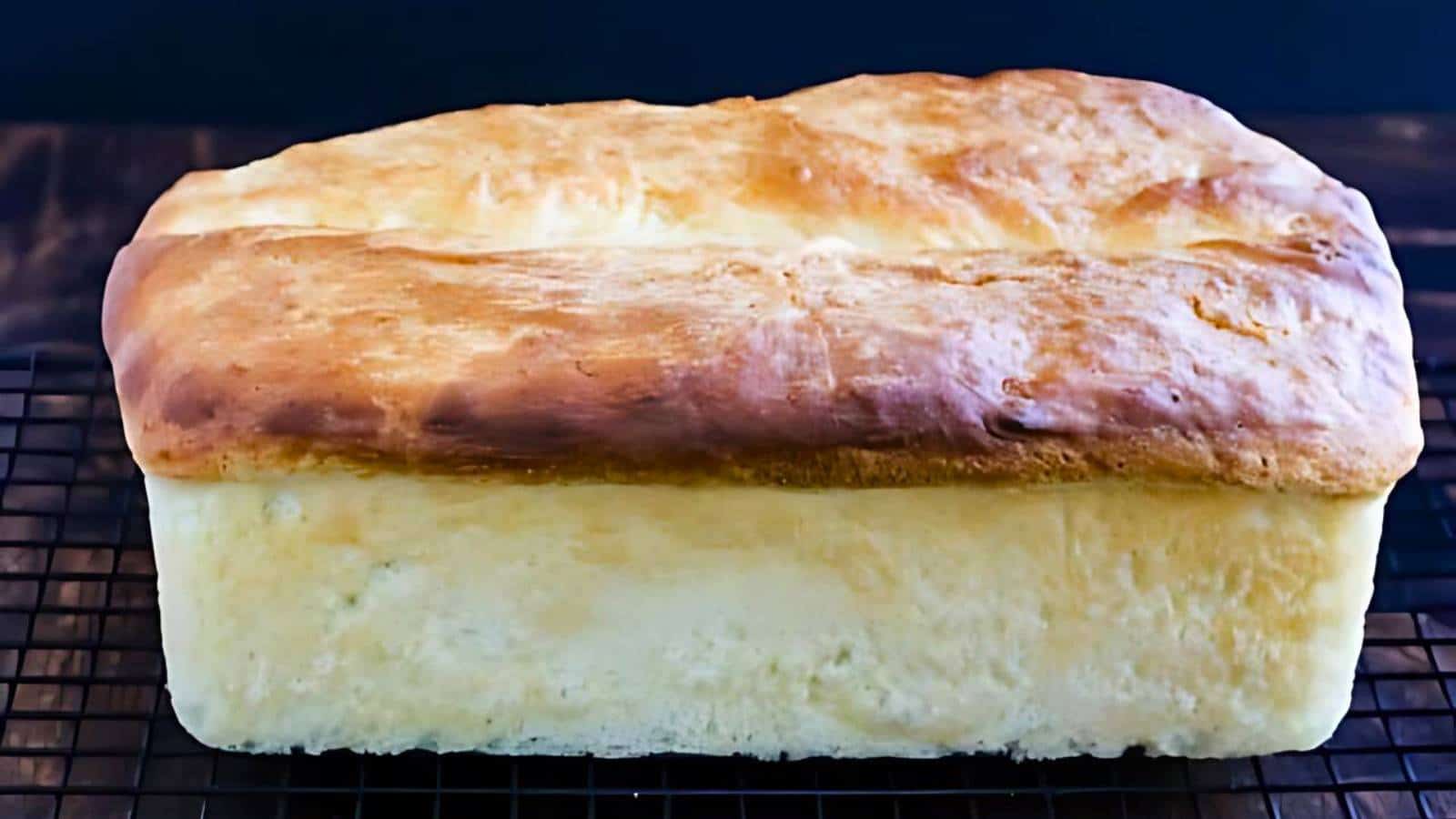 A golden-brown loaf of bread cools on a black wire rack against a dark background, perfect for cozy fall bread baskets.
