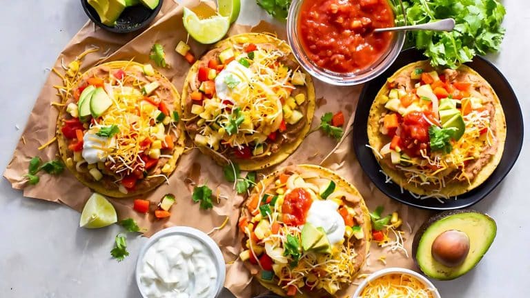 Four loaded tostadas topped with various colorful vegetables, cheese, sour cream, avocado, and salsa on parchment paper. Bowls of salsa, sour cream, and a halved avocado are on the side.