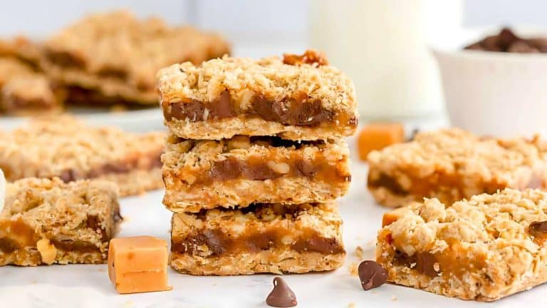 A stack of three caramel oatmeal bars is placed on a white surface, surrounded by additional bars, caramel squares, and chocolate chips. A glass of milk and a bowl of chocolate chips are in the background.