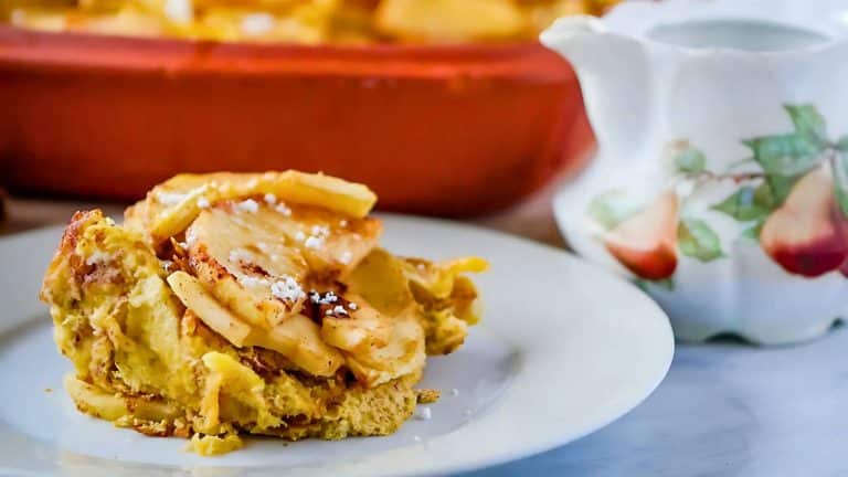 A slice of apple casserole on a white plate, with a floral-patterned pitcher in the background.