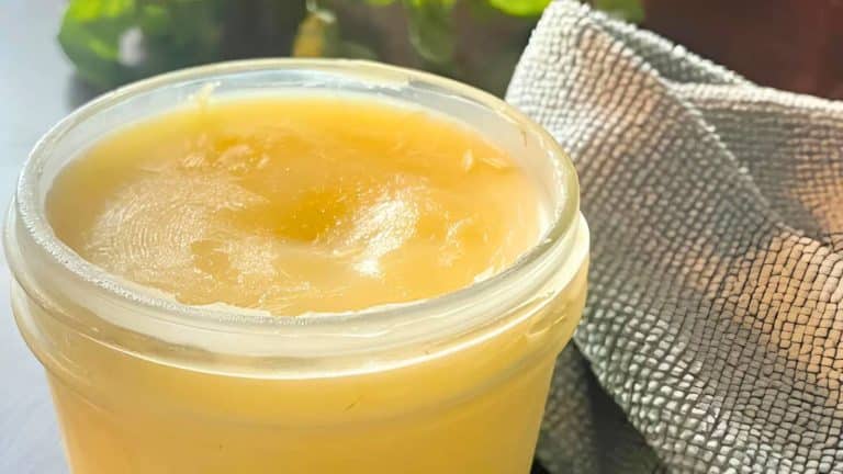 A glass jar filled with creamy, yellow ghee sits next to a textured gray cloth, with green foliage blurred in the background.