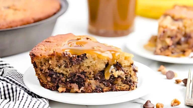A slice of chocolate chip banana bread with caramel drizzle on a white plate, next to a whole loaf in a pan and a glass of drink. A striped cloth is partially visible nearby.