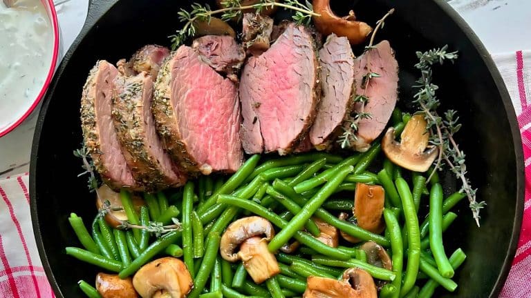 Sliced medium-rare roast beef with herbs, served with green beans and mushrooms in a black skillet.