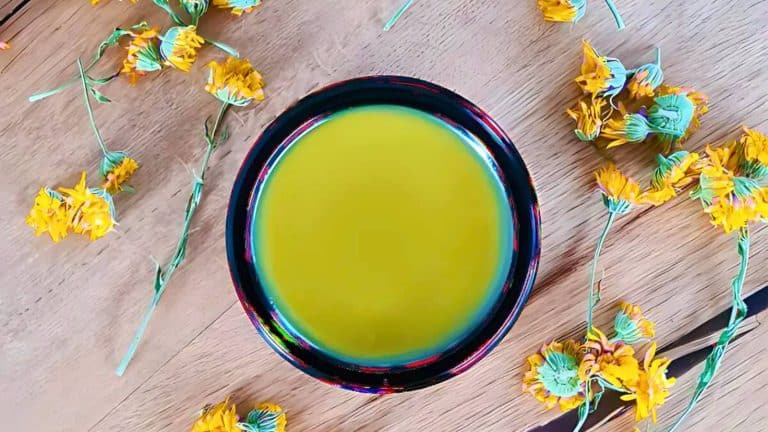 A bowl of yellow liquid on a wooden surface surrounded by scattered yellow flowers.