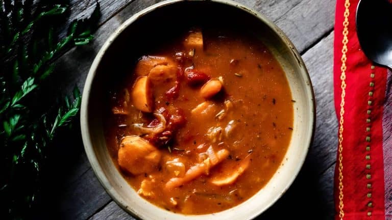 A bowl of hearty tomato-based stew with chunks of meat and vegetables on a wooden surface, next to a red cloth with a gold border.