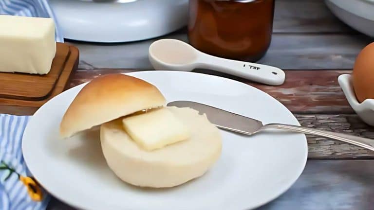 A buttered roll, one of those tasty breads that instantly delights the senses, sits on a plate with a gleaming butter knife. Surrounding it are a polished butter dish, an elegant measuring spoon, and a perfectly placed egg in its holder.