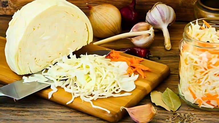Chopped cabbage and carrots on a cutting board with a knife, surrounded by onions, garlic, and spices. A jar of prepared slaw is nearby.