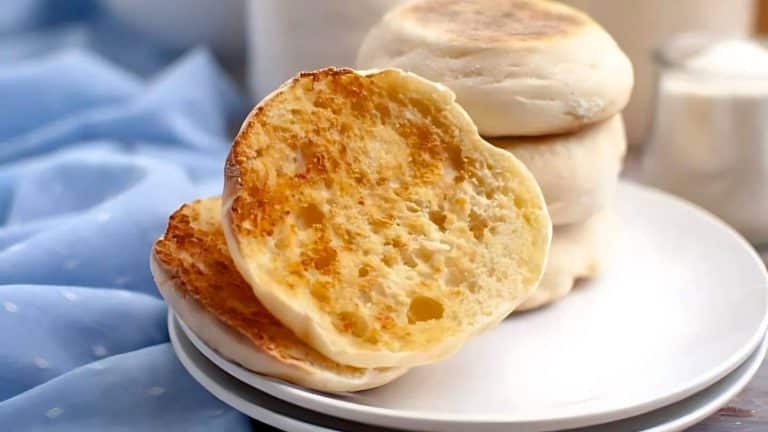 Toasted English muffin halves on a plate with a stack of uncut muffins in the background.