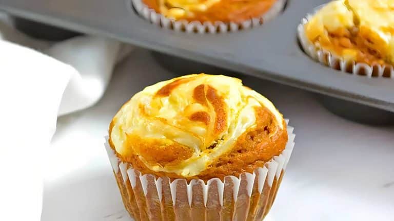 A pumpkin cream cheese muffin sits in a paper liner on a white counter, with more muffins in a metal pan in the background.