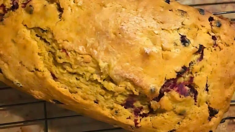 A close-up of a baked loaf of bread with a golden-brown crust, dotted with visible berries, on a cooling rack.