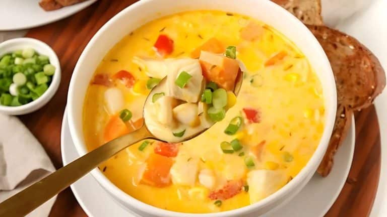 A bowl of creamy soup with corn, diced vegetables, and garnished with chopped green onions. A spoon lifts a portion. Slices of brown bread are on the side.
