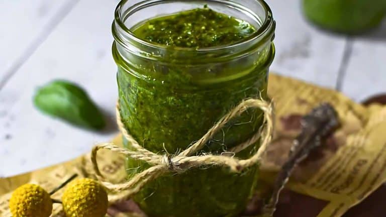 Glass jar filled with green pesto sauce, wrapped with twine, placed on a paper and wooden surface. A basil leaf and yellow flowers are nearby.