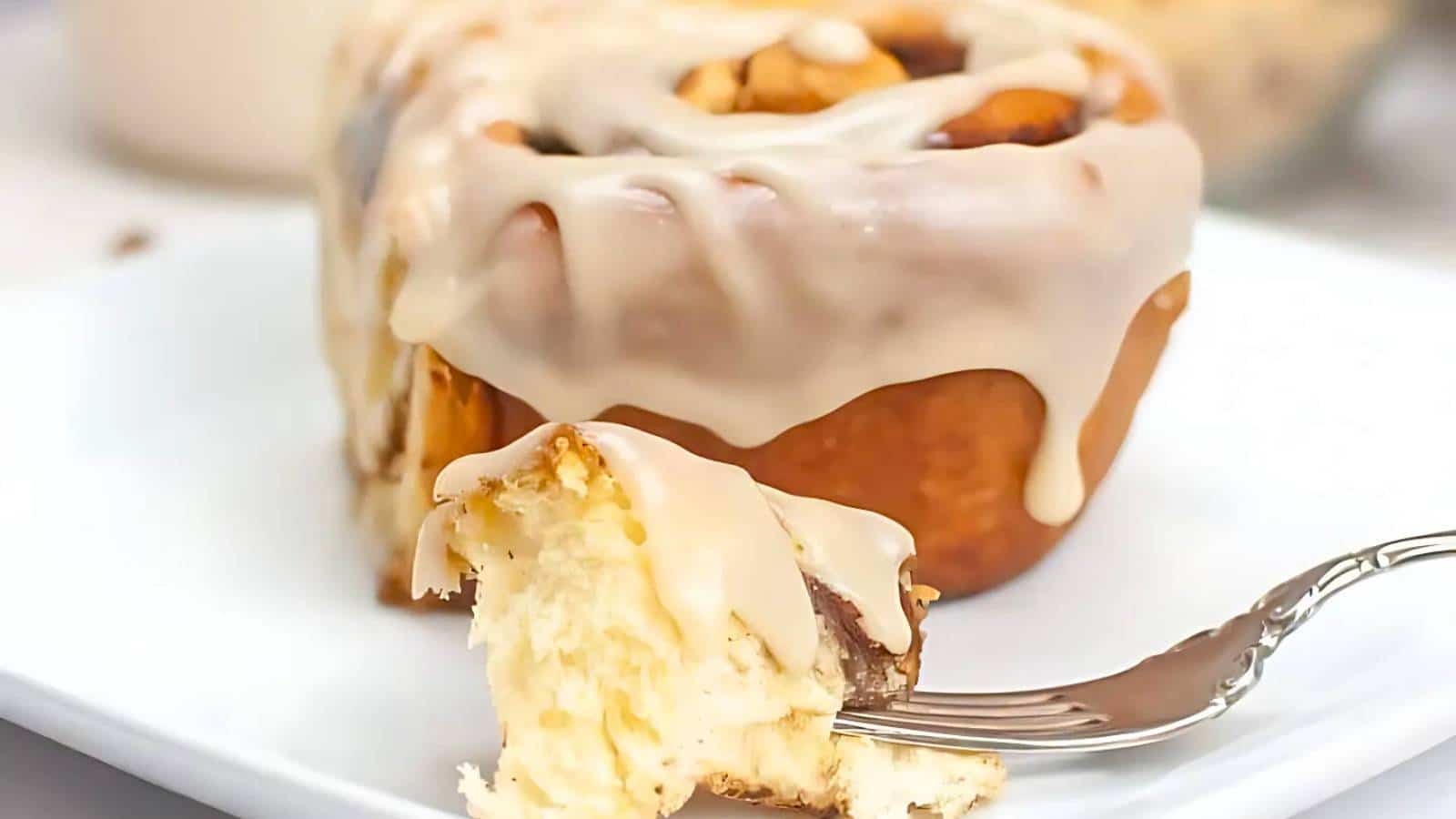 A cinnamon roll with icing on top sits on a white plate. A fork has taken a piece from the roll.