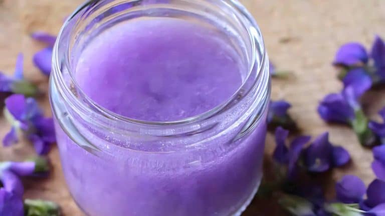 A jar filled with purple gel sits on a wooden surface, surrounded by scattered small purple flowers.