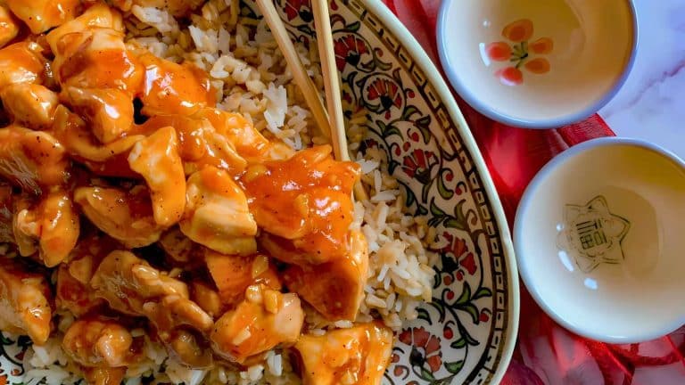 A decorative plate with chicken in orange sauce over rice, two empty bowls beside it, and wooden chopsticks on a red cloth.