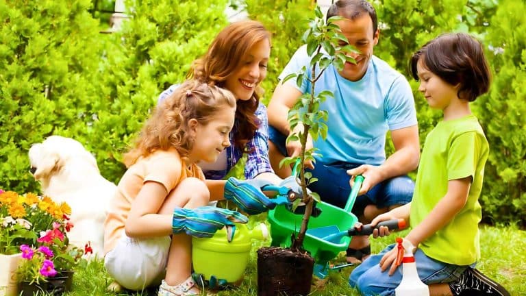 A family of four, including a mother, father, and two children, planting a small tree together in a garden. The children are using green watering cans and hand shovels, and a dog is in the background.