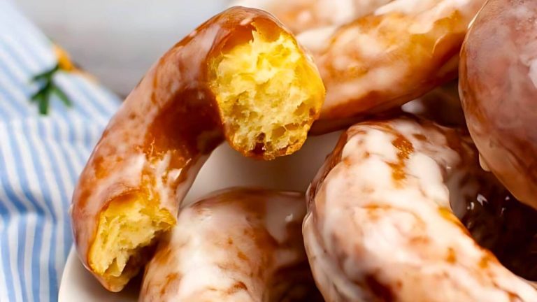 Close-up of glazed donuts stacked on a plate, with one donut partially eaten, revealing its fluffy interior. A blue-striped cloth is partially visible in the background.