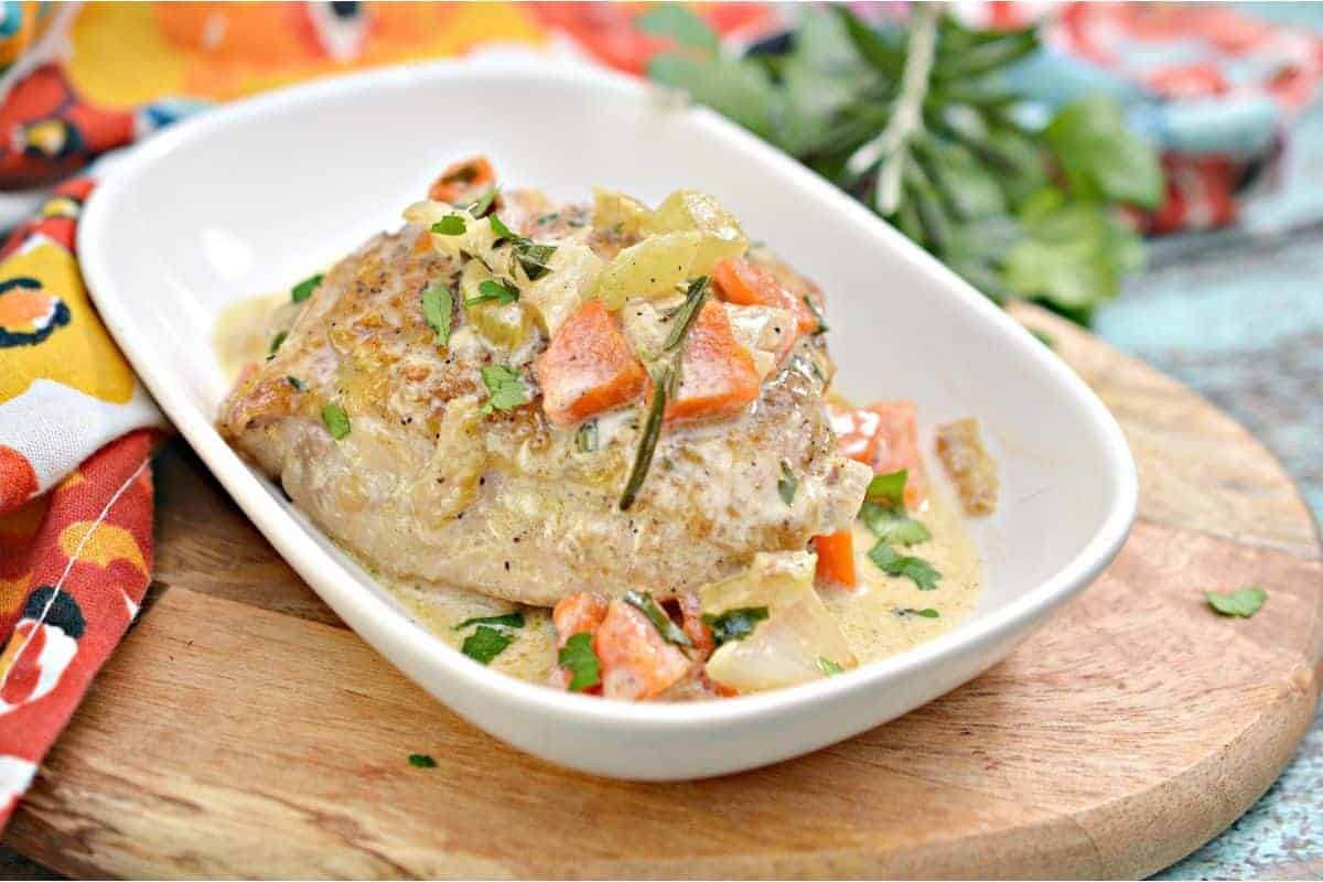 Plate of cooked pork chop topped with carrots, herbs, and creamy sauce on a white dish, resting on a wooden board with colorful fabric in the background.