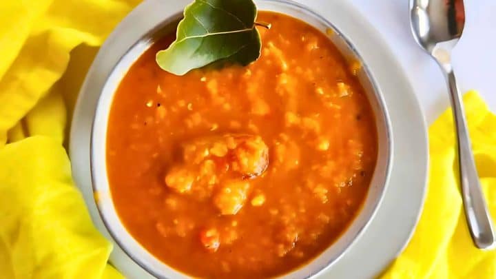 A bowl of orange lentil soup garnished with a bay leaf is placed on a white table with a silver spoon nearby.