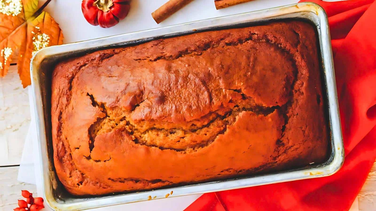 A freshly baked loaf of pumpkin bread in a rectangular pan, surrounded by a red cloth, autumn leaves, a small pumpkin, and a cinnamon stick.