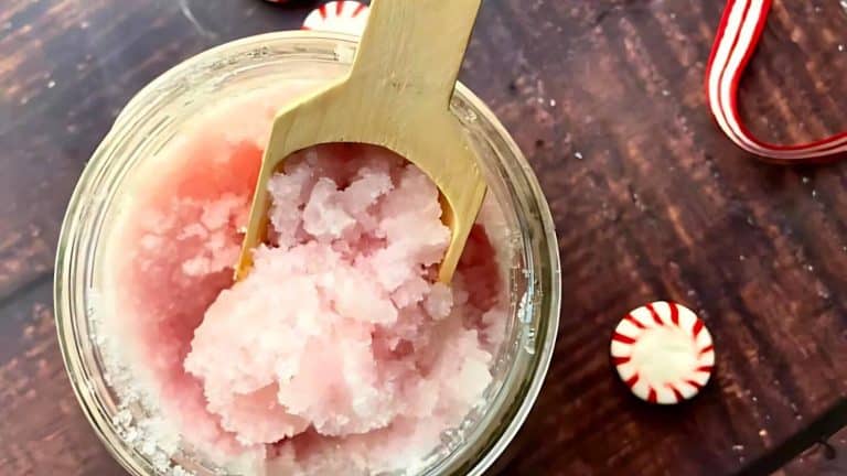 A glass jar of pink sugar scrub with a wooden spoon on a wooden surface, next to a peppermint candy and a red and white ribbon.