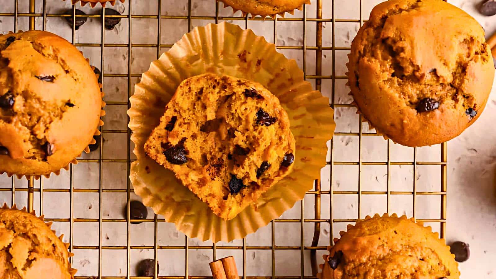 Pumpkin chocolate chip muffins on a wire rack, with one muffin partially unwrapped and cut in half to show the inside.