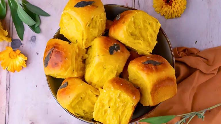 A bowl of golden-brown bread rolls with some leaves on top, placed on a table with flowers and a brown cloth nearby.