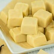 A white plate with cubes of creamy vanilla fudge on parchment paper, next to a few vanilla pods.