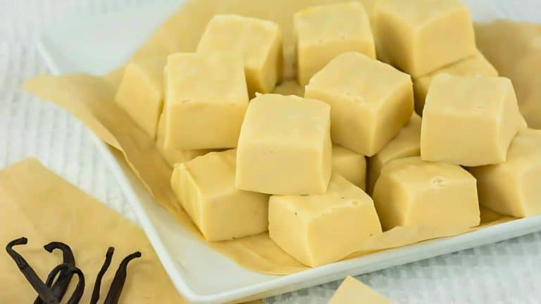 A white plate with cubes of creamy vanilla fudge on parchment paper, next to a few vanilla pods.