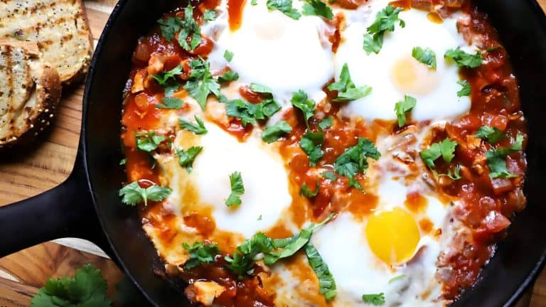 A skillet with shakshuka, featuring poached eggs in a spiced tomato sauce, garnished with cilantro. Toasted bread slices are on the side.