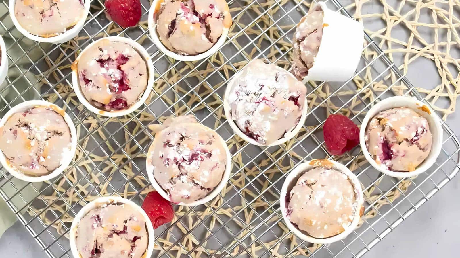 A cooling rack with multiple muffins in white paper cups, featuring a raspberry filling. Fresh raspberries are scattered around.