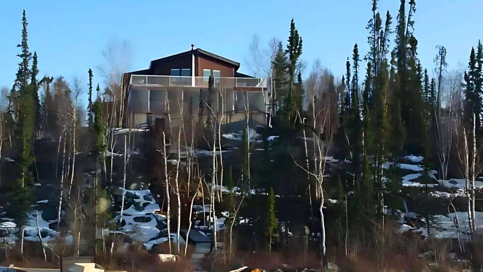House atop a wooded, snowy hill with tall trees, seen from below on a clear day.