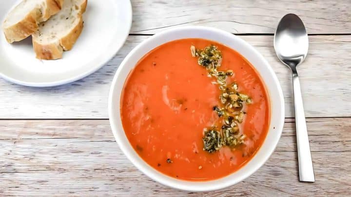 A bowl of tomato soup garnished with herbs and seeds, next to a spoon and a plate with slices of bread on a wooden table.