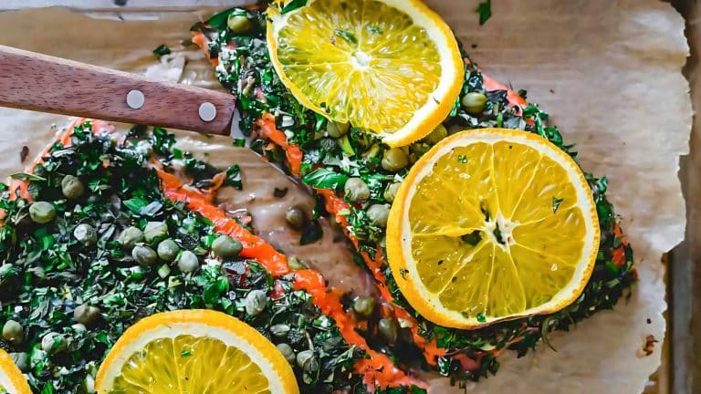 Salmon fillet garnished with sliced oranges, capers, and chopped herbs on parchment paper. A wooden-handled knife rests on the fillet.