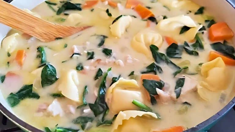 Soup with tortellini, spinach, chicken, and carrots in a creamy broth, being stirred with a wooden spoon.