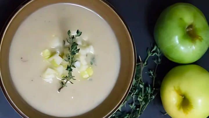 A bowl of creamy soup garnished with thyme and diced apples, next to two whole green apples on a dark surface.