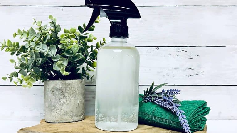 Clear spray bottle on a wooden surface, next to a small potted plant and a green folded cloth with lavender sprigs. Background features white wooden panels.