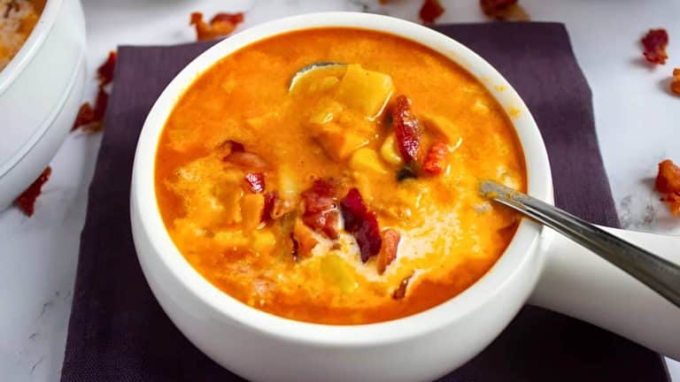 A white bowl filled with creamy, orange-hued soup, topped with crispy bacon bits, placed on a dark napkin. A spoon is partially submerged in the soup.