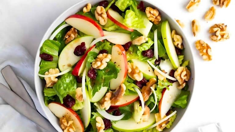 A salad in a bowl with apple slices, walnuts, cranberries, and lettuce. A few walnuts are scattered beside the bowl.