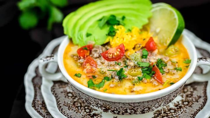 A bowl of creamy soup topped with slices of avocado, chopped tomatoes, grated cheese, cilantro, and a lime wedge on a decorative plate.