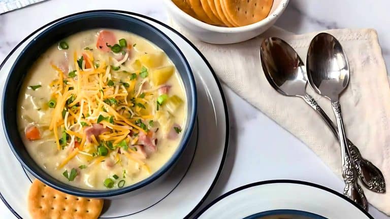 A bowl of creamy potato and ham soup topped with shredded cheese and green onions. Crackers and two spoons are placed beside the dish.